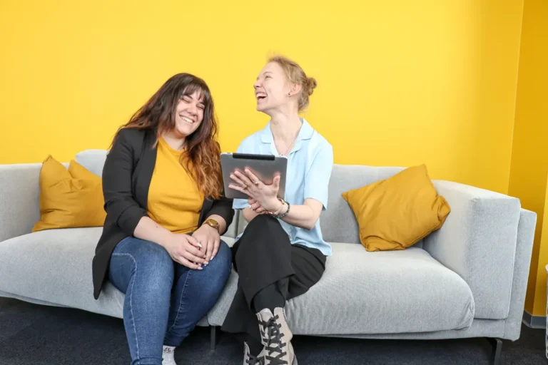 Marketing Managerin Sarah Gern und Redakteurin Maleen Thiele von VTH Marketing & Media lachend und auf ein Tablet schauend im VTH-Kreativraum.
